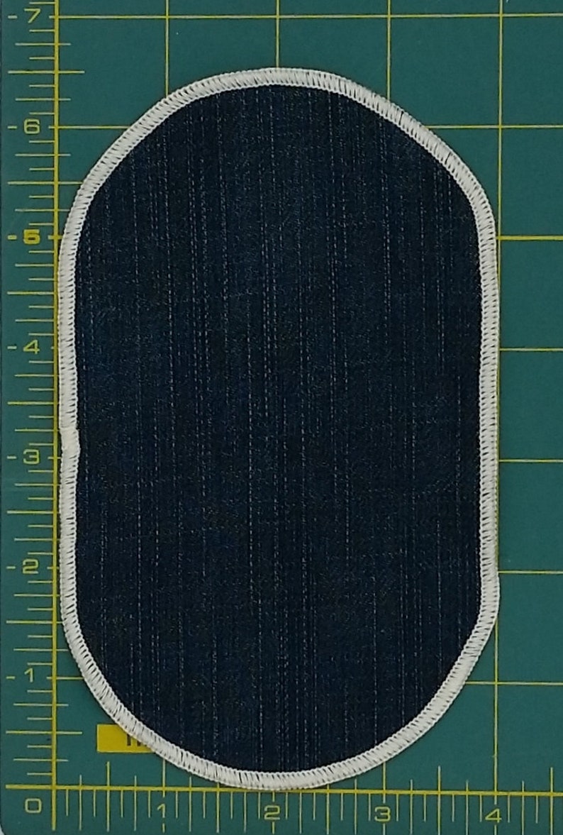 May include: Oval-shaped patch with a dark blue, vertically striped fabric center. The patch is outlined with a white, stitched border. The background is a green cutting mat with measurement markings.