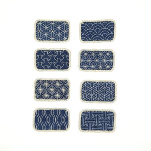Könnte beinhalten: Acht blaue Stoffpatches mit weißen, gestickten geometrischen Mustern. Die Patches sind rechteckig und haben einen cremefarbenen Rand.