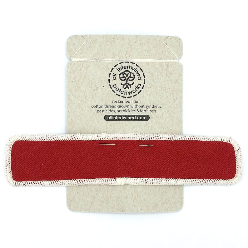 May include: A red fabric patch with white stitching around the edges. The patch is on a brown cardboard backing with the text "all intertwined patchwork reclaimed fabric cotton thread grown without synthetic pesticides, herbicides & fertilizers allintertwined.com".