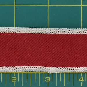 May include: A red fabric name tag with white stitching around the edges. The tag is approximately 12.7 cm long and 2.5 cm wide.