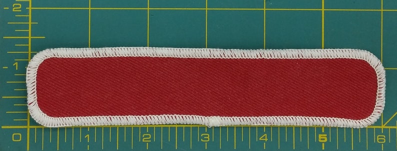 May include: A red fabric name tag with white stitching around the edges. The tag is approximately 12.7 cm long and 2.5 cm wide.