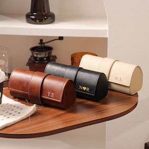 Może przedstawiać: Trzy skórzane etui na zegarki w kolorze brązowym, czarnym i kremowym, każde z personalizowaną inicjałem. Na brązowym etui nadrukowano "T.E", na czarnym etui "N♥R", a na kremowym etui "W.K". Etui są cylindryczne z zapięciem na pasek.