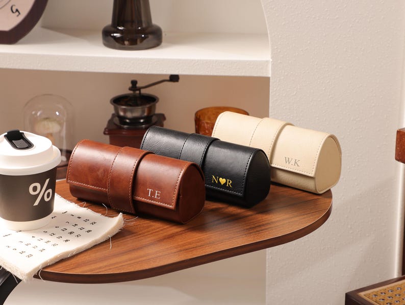 May include: Three leather watch cases in brown, black, and cream colors, each with a personalized initial. The brown case has "T.E" printed on it, the black case has "N&hearts;R", and the cream case has "W.K". The cases are cylindrical with a strap closure.