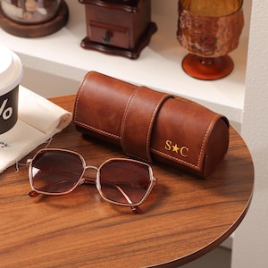 May include: A pair of rose-tinted sunglasses with a gold-colored frame rests on a wooden surface next to a brown leather glasses case. The case is embossed with the initials "S*C". A coffee cup with the "%" symbol is also visible.