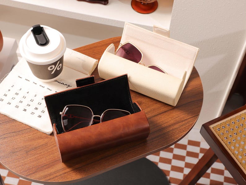 May include: A brown leather glasses case with a pair of sunglasses inside, next to a white glasses case with sunglasses. A coffee cup with the percent symbol is on a calendar.