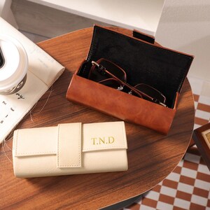 May include: Two leather eyeglass cases, one open with glasses inside. The open case is brown, the closed case is cream-colored with "T.N.D" in gold lettering. A white travel mug and a calendar are also on the wooden surface.