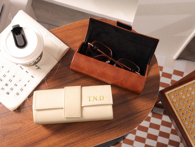May include: Two leather eyeglass cases, one open with glasses inside. The open case is brown, the closed case is cream-colored with "T.N.D" in gold lettering. A white travel mug and a calendar are also on the wooden surface.