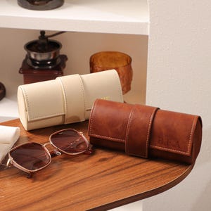 May include: A pair of brown-tinted sunglasses with a gold frame rests on a wooden surface, next to a beige and a brown leather watch roll. The brown watch roll has initials engraved on it. Other items are visible in the background.