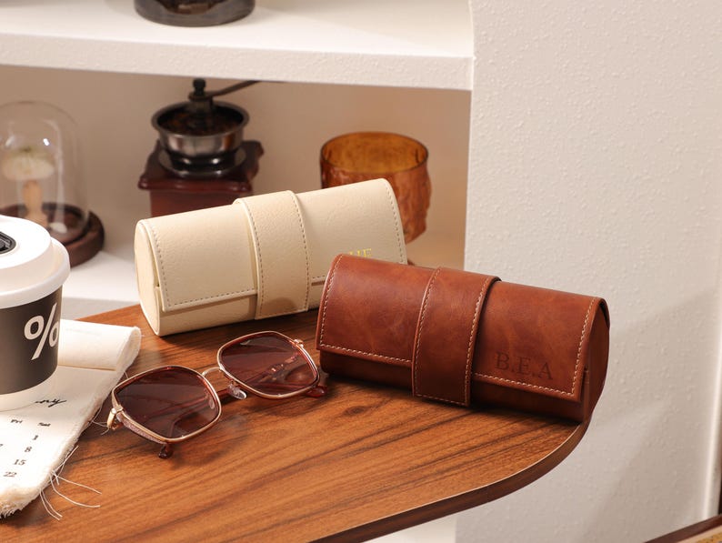 May include: A pair of brown-tinted sunglasses with a gold frame rests on a wooden surface, next to a beige and a brown leather watch roll. The brown watch roll has initials engraved on it. Other items are visible in the background.