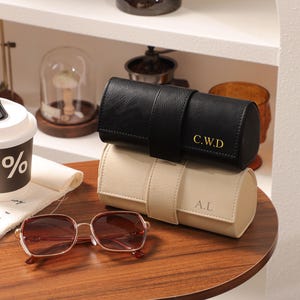 May include: Two leather watch cases, one black and one beige, stacked on a wooden surface. The black case has gold initials "C.W.D", and the beige case has "A.L". A pair of brown-tinted sunglasses and a coffee cup with a "%" symbol are also visible.
