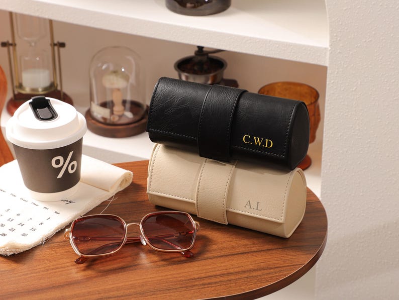 May include: Two leather watch cases, one black and one beige, stacked on a wooden surface. The black case has gold initials "C.W.D", and the beige case has "A.L". A pair of brown-tinted sunglasses and a coffee cup with a "%" symbol are also visible.
