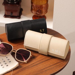 May include: A pair of sunglasses with a gold frame and pink lenses rests on a wooden table next to a black and a cream-colored leather eyeglass case. The black case has the initials "S.T" and the cream case has the initials "A.M.S."