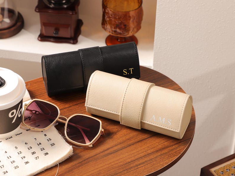 May include: A pair of sunglasses with a gold frame and pink lenses rests on a wooden table next to a black and a cream-colored leather eyeglass case. The black case has the initials "S.T" and the cream case has the initials "A.M.S."