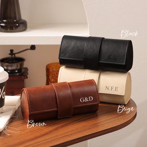 May include: Three leather watch cases in black, beige, and brown, each with a strap closure. The black case has the initials "K.S", the beige case has "N.F.E", and the brown case has "G&D". The cases are cylindrical and designed for storing watches.