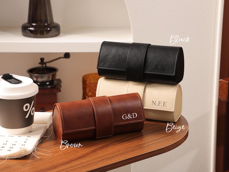 May include: Three leather watch cases in black, beige, and brown, each with a strap closure. The black case has the initials "K.S", the beige case has "N.F.E", and the brown case has "G&D". The cases are cylindrical and designed for storing watches.