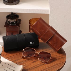 May include: A pair of square-framed sunglasses with a gold-colored frame and brown lenses rests on a wooden surface. Beside them are two leather eyeglass cases, one black and one brown, each with initials. A coffee cup and calendar are in the foreground.