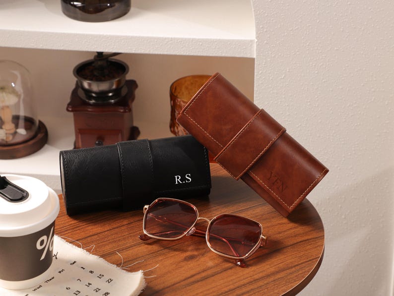 May include: A pair of square-framed sunglasses with a gold-colored frame and brown lenses rests on a wooden surface. Beside them are two leather eyeglass cases, one black and one brown, each with initials. A coffee cup and calendar are in the foreground.