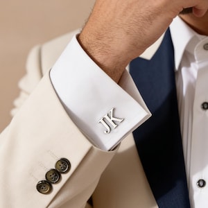 Könnte beinhalten: Nahaufnahme eines cremefarbenen Anzugs mit marineblauem Revers und weißem Hemd. Der Manschettenknopf zeigt die Initialen "JK" in Silber. Die Jacke hat drei dunkelbraune Knöpfe.