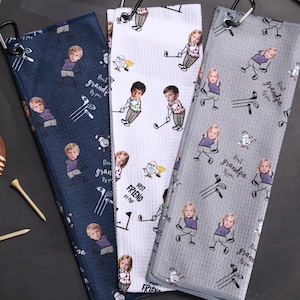 May include: Three golf towels with cartoon illustrations of people and golf clubs. One towel is navy blue, one is white, and one is gray. Each towel has a black carabiner clip attached. The words "Best Friend" and "Best Grandpa" are printed on the towels.