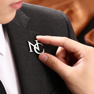 Op de afbeelding: Een zilverkleurige reversspeld met de initialen "NC" wordt op een donkergrijze blazer geplaatst. De persoon draagt een wit overhemd en een zwarte stropdas. Een glas amberkleurige vloeistof staat op een houten dienblad op de achtergrond.