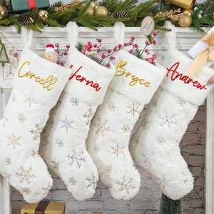 May include: Four white faux fur Christmas stockings with snowflake designs and personalized names in gold and red embroidery. The names are "Corecll", "Jenna", "Bryce", and "Andrew".