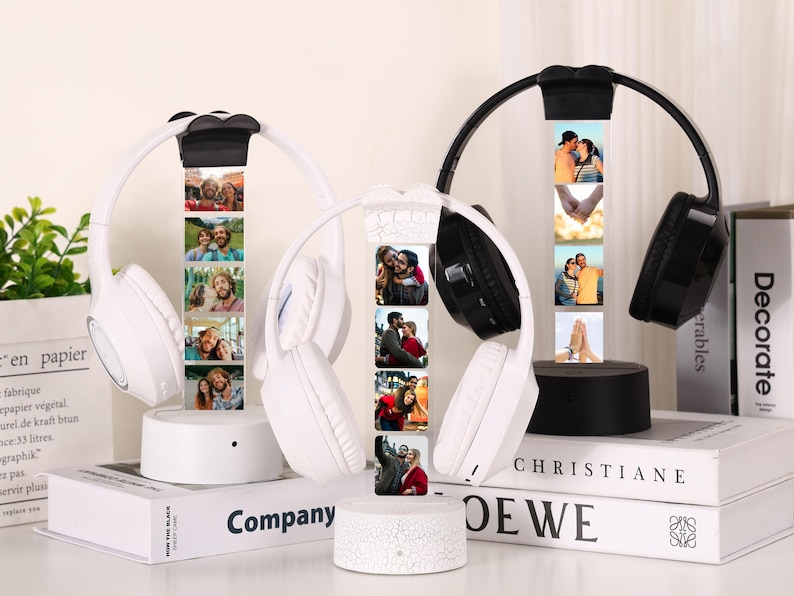 Pu&ograve; includere: Cuffie wireless bianche e nere con display fotografico. Le cuffie sono su una base bianca, con una serie di foto visualizzate verticalmente. Le cuffie poggiano su libri con le parole "Company" e "Decorate" visibili.