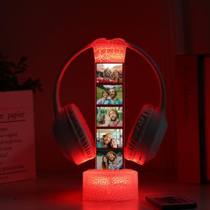 Pu&ograve; includere: Una lampada decorativa illuminata di rosso con un paio di cuffie bianche e una striscia verticale di foto. La base e la parte superiore della lampada hanno un effetto vetro incrinato. La lampada &egrave; su un tavolo con libri e un sacchetto di carta.