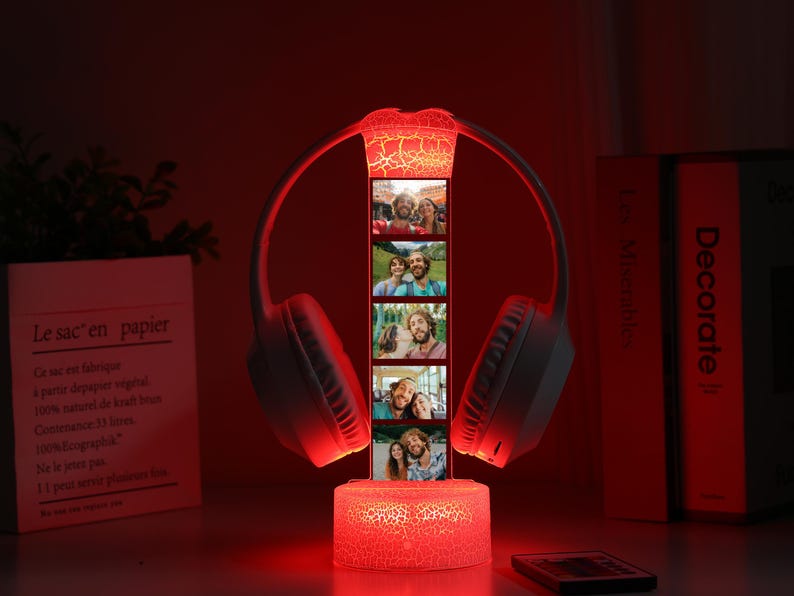 Pu&ograve; includere: Una lampada decorativa illuminata di rosso con un paio di cuffie bianche e una striscia verticale di foto. La base e la parte superiore della lampada hanno un effetto vetro incrinato. La lampada &egrave; su un tavolo con libri e un sacchetto di carta.