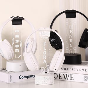 May include: White and black wireless headphones displayed on illuminated stands. The stands feature names and decorative designs. The headphones rest on books, with the names "MASON", "FLORA", and "HENRY" visible.