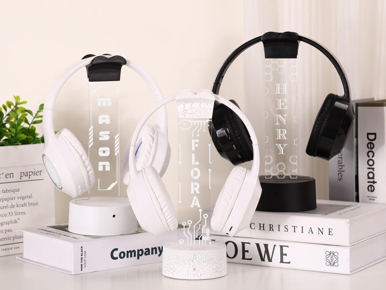 May include: White and black wireless headphones displayed on illuminated stands. The stands feature names and decorative designs. The headphones rest on books, with the names "MASON", "FLORA", and "HENRY" visible.