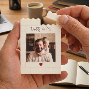 May include: A white rectangular building block frame featuring the text Daddy & Me in black script, a small red heart icon, and a central photograph of an adult and a child. The frame is constructed from interlocking plastic bricks.