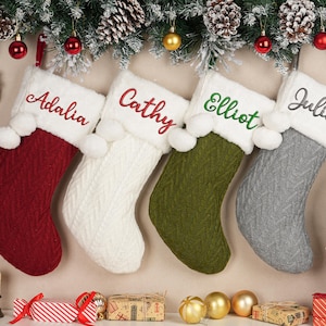 Peut inclure: Quatre bas de Noël personnalisés, rouges, blancs, verts et gris, chacun brodé d'un nom de couleur différente. Les bas sont suspendus à une guirlande de pin et d'ornements, créant une scène de vacances festive.