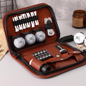 Op de afbeelding: Een bruine leren golfset met ritssluiting, met golfballen, tees, een divotgereedschap en een scorekaarthouder. De set bevat drie golfballen met een logo en een zwart-wit ruitpatroon.