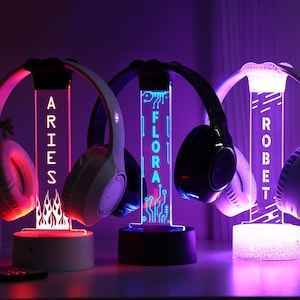May include: Three illuminated headphone stands with personalized names: ARIES, FLORA, and ROBET. Each stand features a unique design and color scheme, with headphones resting on top. The stands glow with vibrant colors, creating a modern aesthetic.