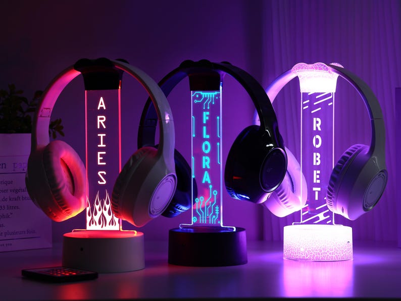 May include: Three illuminated headphone stands with personalized names: ARIES, FLORA, and ROBET. Each stand features a unique design and color scheme, with headphones resting on top. The stands glow with vibrant colors, creating a modern aesthetic.
