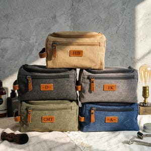 Puede incluir: Un conjunto de seis neceseres de lona en varios colores: beige, gris, verde oliva y azul. Cada bolsa tiene una cremallera, detalles de cuero y un parche de cuero personalizado con iniciales. Las bolsas están diseñadas para viajar.