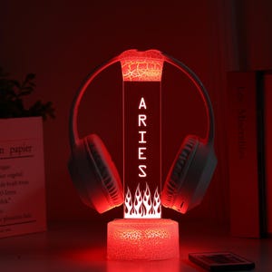 May include: A red LED lamp with the word "ARIES" vertically displayed, flanked by a pair of white headphones. The lamp has a flame design at the base and a cracked glass effect. A remote control and books are in the background.