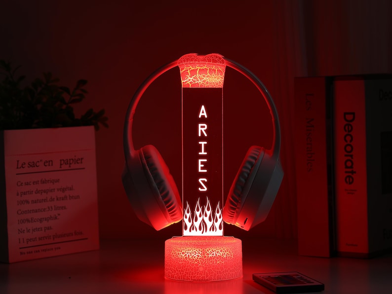 May include: A red LED lamp with the word "ARIES" vertically displayed, flanked by a pair of white headphones. The lamp has a flame design at the base and a cracked glass effect. A remote control and books are in the background.