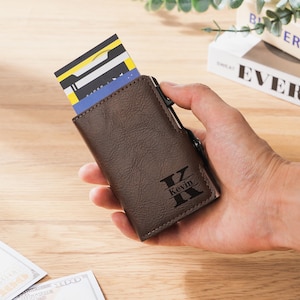 May include: A brown leather wallet with a personalized initial and name, holding several credit cards. The wallet is rectangular and has a textured surface. The cards are visible at the top of the wallet. The wallet is held in a hand.