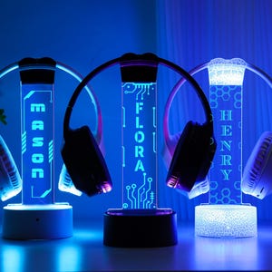 May include: Three illuminated headphone stands with personalized names: MASON, FLORA, and HENRY. Each stand features a vertical light-up panel with a name, a base, and a curved top to hold headphones. The stands emit a blue glow.