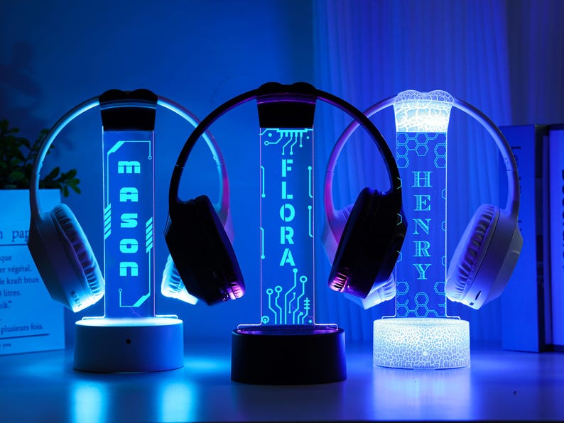 May include: Three illuminated headphone stands with personalized names: MASON, FLORA, and HENRY. Each stand features a vertical light-up panel with a name, a base, and a curved top to hold headphones. The stands emit a blue glow.