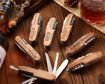 Puede incluir: Navajas multiusos de madera grabadas. Cada herramienta presenta un nombre y una fecha, con frases como "Grooms Man" y "Best Man". Las navajas están dispuestas sobre una superficie de madera, junto a una botella y un vaso de líquido ámbar.
