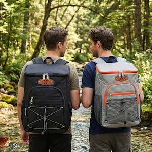 May include: Two insulated backpacks, one black, one grey with orange trim. Both have multiple pockets and bungee cord detailing. The black backpack has a brown leather patch. Ideal for picnics or outdoor adventures.