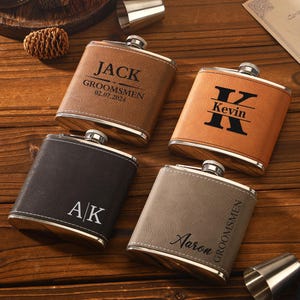 May include: Four personalized leather flasks in shades of brown, tan, and gray, each with a silver cap. The flasks are engraved with names and initials, including "Jack," "Kevin," "AK," and "Aaron." The flasks are arranged on a wooden surface.