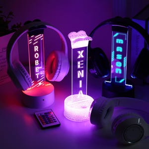 May include: Three illuminated headphone stands with personalized names, each with a pair of headphones. The stands glow with red, blue, and purple lights. A remote control is in the foreground. The names "ROBERT", "XENIA", and "ERROC" are displayed.