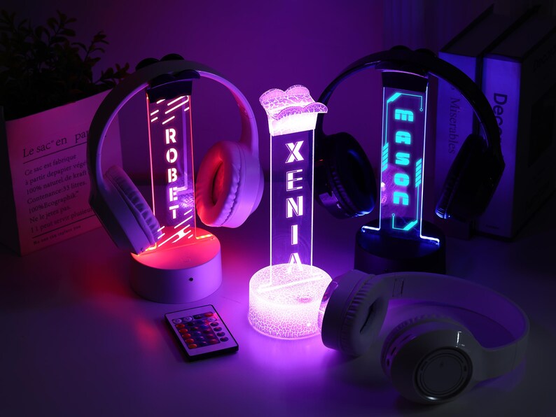 May include: Three illuminated headphone stands with personalized names, each with a pair of headphones. The stands glow with red, blue, and purple lights. A remote control is in the foreground. The names "ROBERT", "XENIA", and "ERROC" are displayed.