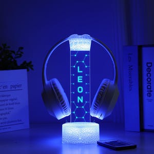 May include: A glowing blue LED headphone stand with the name "LEON" illuminated. The stand holds white headphones and sits on a table with a remote and a decorative plant in a white paper pot. The background includes books.