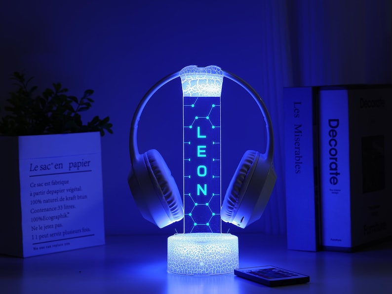 May include: A glowing blue LED headphone stand with the name "LEON" illuminated. The stand holds white headphones and sits on a table with a remote and a decorative plant in a white paper pot. The background includes books.