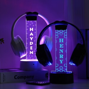 May include: Two illuminated headphone stands with the names "HAYDEN" and "HENRY" in white and blue. Each stand holds a pair of headphones. The stands have a geometric design and are lit with purple and blue lights.