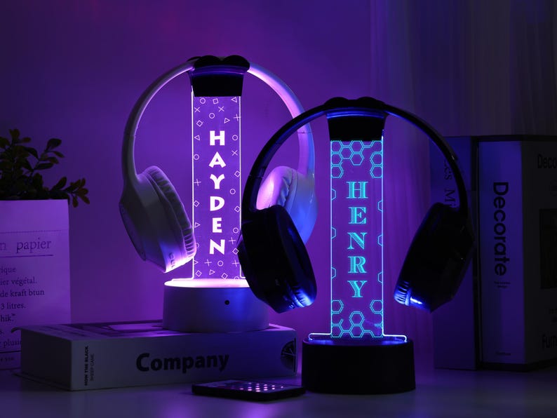 May include: Two illuminated headphone stands with the names "HAYDEN" and "HENRY" in white and blue. Each stand holds a pair of headphones. The stands have a geometric design and are lit with purple and blue lights.
